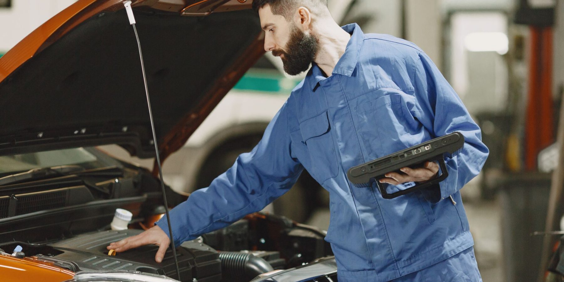 Kfz-Mechaniker im blauen Overall prüft mit einem Diagnosegerät den Motorraum eines Fahrzeugs